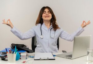 with closed eyes smiling middle aged female doctor wearing wearing medical robe with stethoscope sitting desk work laptop with medical tools showing okey gesture white wall 141793 23552